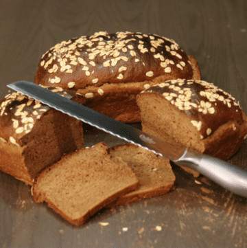 two loaves of homemade brown pumpernickel bread with rolled oats on top, one loaf is whole and the other loaf is sliced in half with 2 slices of bread showing with a metal bread knife leaning on one half of the sliced bread