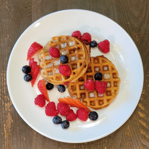Beautiful picture of a white plate and two spelt waffles, with melted butter, syrup, and fresh berries. Strawberries, blueberries, and red raspberries sprinkled all over the plate and both waffles