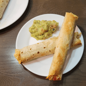 two baked chicken flautas crossed on a white plate with fresh guacamole on the side of the plate