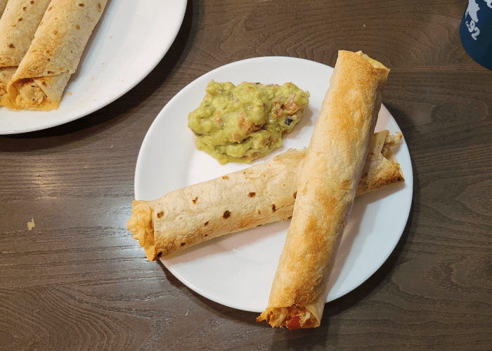 two baked chicken flautas crossed on a white plate with fresh guacamole on the side of the plate