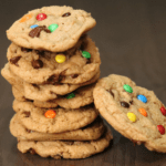 a stack of 7 baked sourdough cookies with another cookie leaning against the stack, m and m candies and chocolate chips are mixed in the cookies