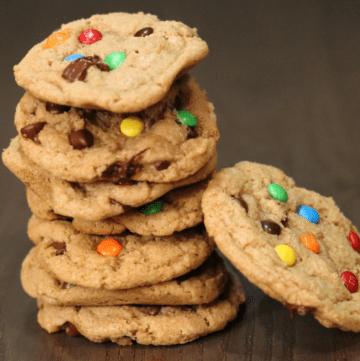 a stack of 7 baked sourdough cookies with another cookie leaning against the stack, m and m candies and chocolate chips are mixed in the cookies