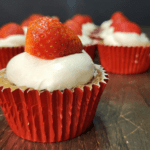 close up of a strawberry sourdough discard cupcake in a red foil cupcake liner with vanilla buttercream on top and garnished with a fresh strawberry, other cupcakes the same way in the background