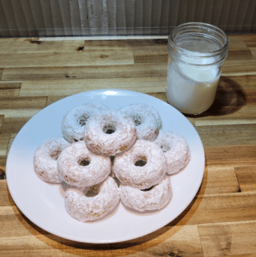 a white plate of white powdered sugar donuts stacked next to a glass jar half full of milk. Both sitting on a wooden countertop