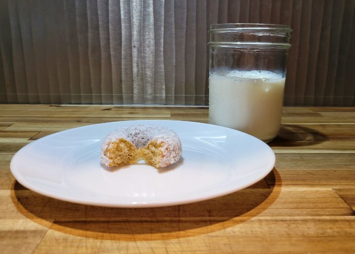 a white plate with a single powdered sugar donut with a bite out of it, sitting next to a glass of milk