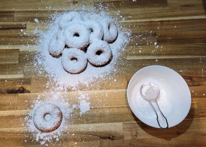 wooden counter with a pile of donuts and a single donut pulled out of the pile, a white bowl filled with powdered sugar and a dusting spoon in the bowl