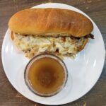 French Dip Sub Hoagie Sandwich sitting on a white plate with a glass cup of Au Jus