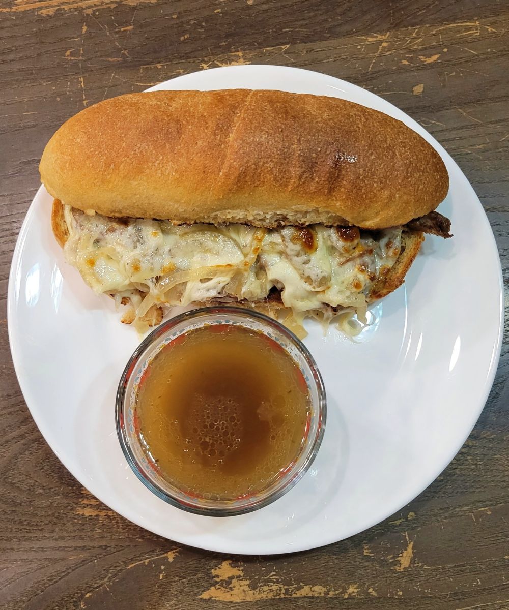 French Dip Sub Hoagie Sandwich sitting on a white plate with a glass cup of Au Jus