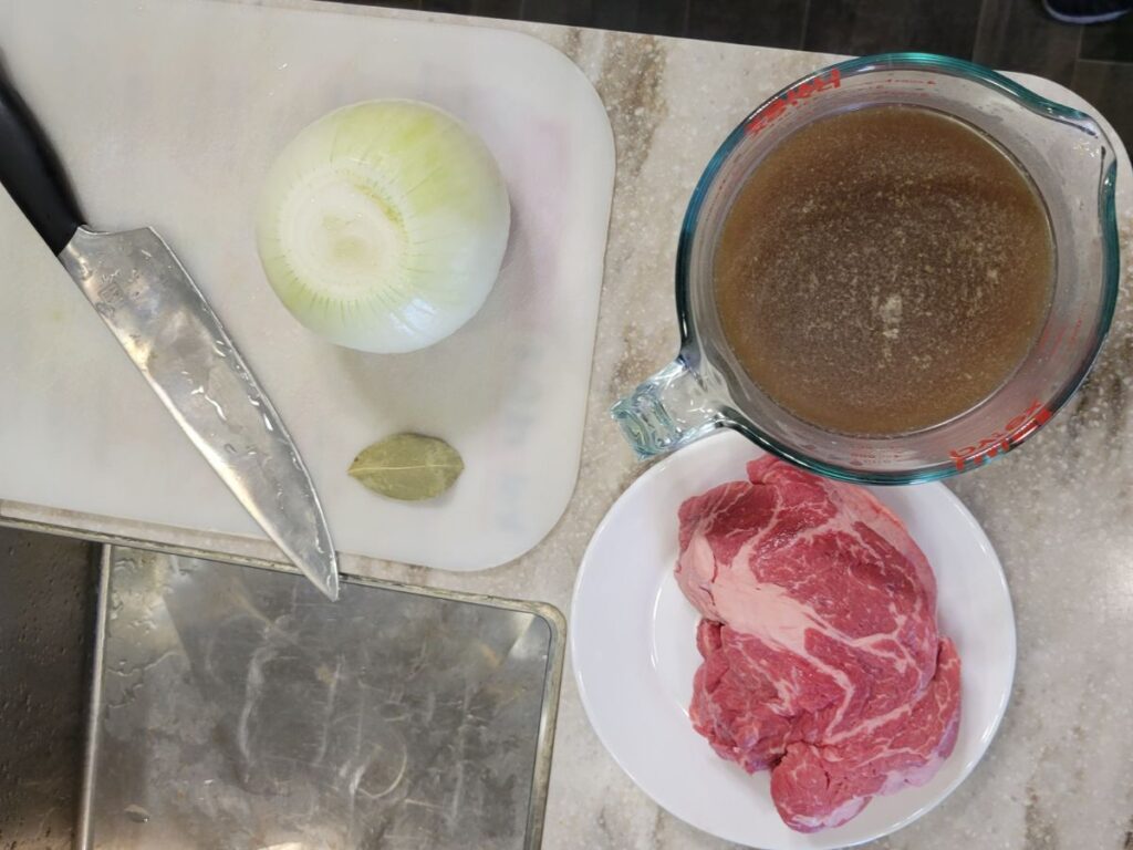 Ingredients to make French Dip Subs from scratch, raw beef chuck roast, broth, onion, and bay leaf, cutting board an knife.