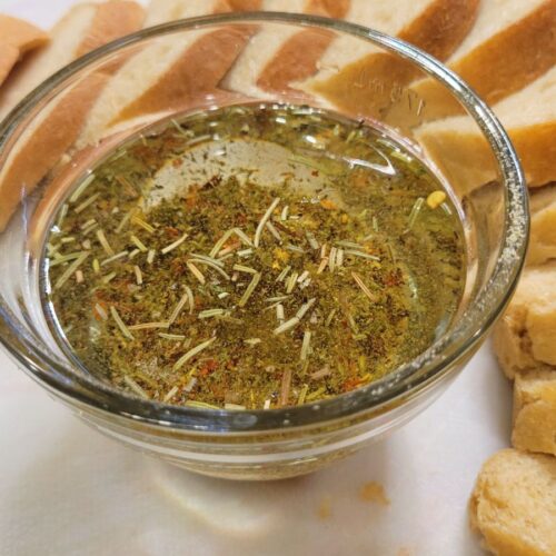 a glass bowl of Simple Dipping Oil with seasonings, and a sliced loaf of bread around the oil.