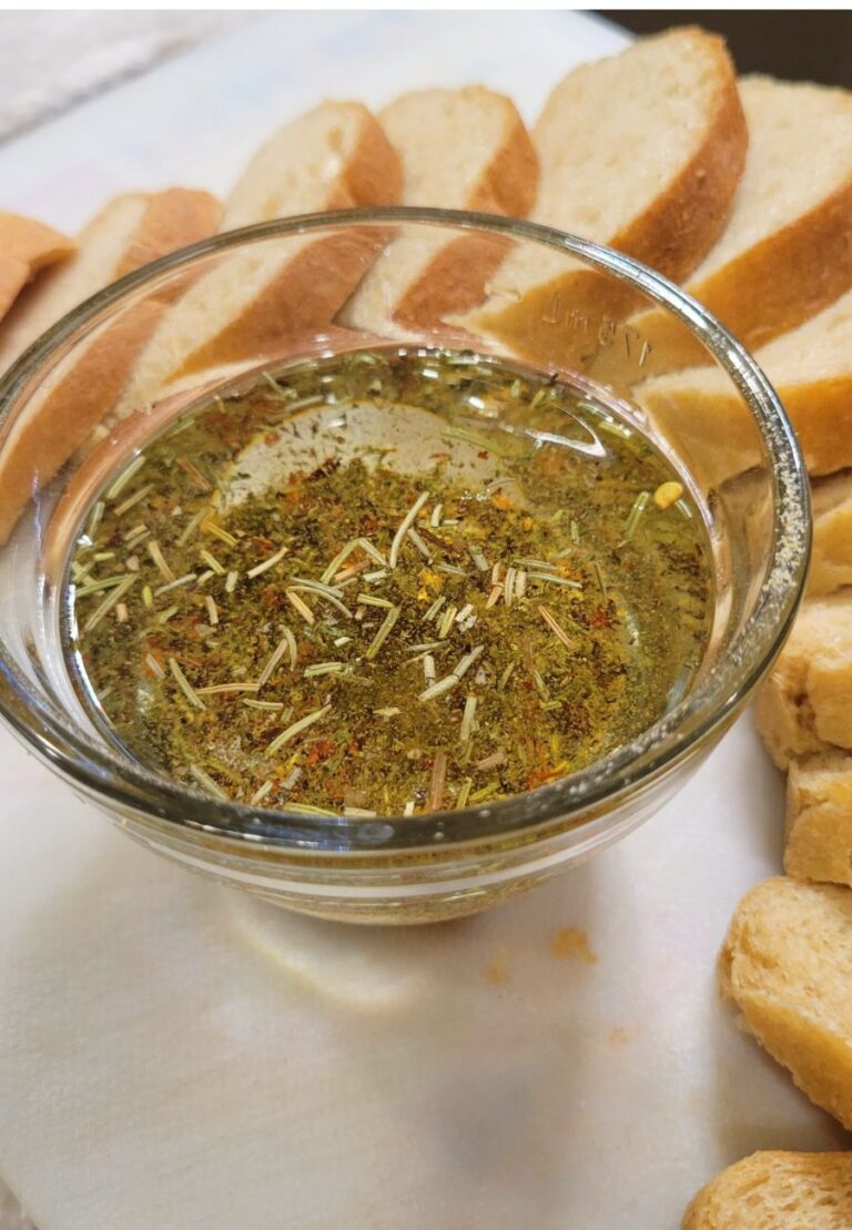 a glass bowl of Simple Dipping Oil with seasonings, and a sliced loaf of bread around the oil.