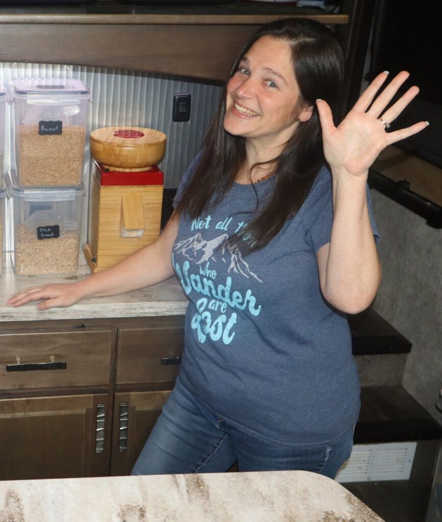 woman standing and waving in front of a flour grain mill and whole grain wheat berries.