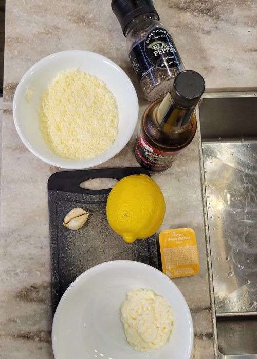 Ingredients for recipe. Lemon, cutting board, parmesan cheese, amber bottle, black pepper, mayo, and garlic