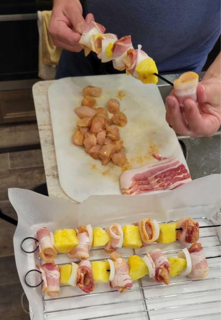 hands skewering on raw chicken, bacon, pineapple, and onions onto metal skewers to make kabobs