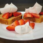a white plate with two slices of pound cake topped with whipped cream and fresh sliced strawberries