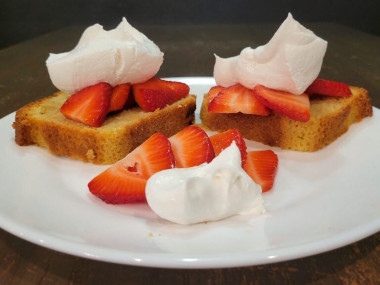 a white plate with two slices of pound cake topped with whipped cream and fresh sliced strawberries