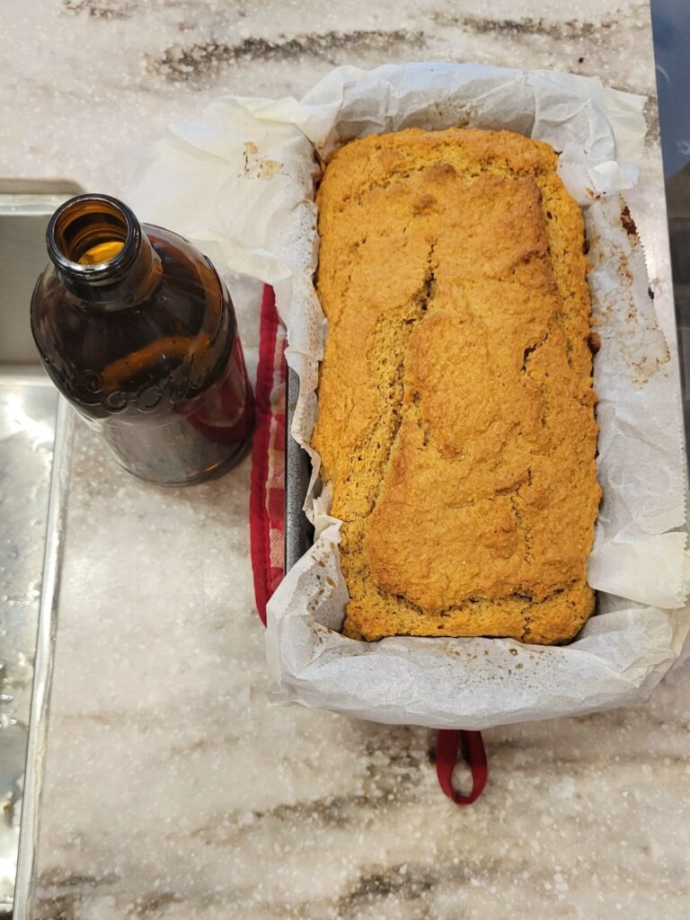 an empty bottle of beer next to a baked loaf of fresh milled flour beer bread in a bread tin.