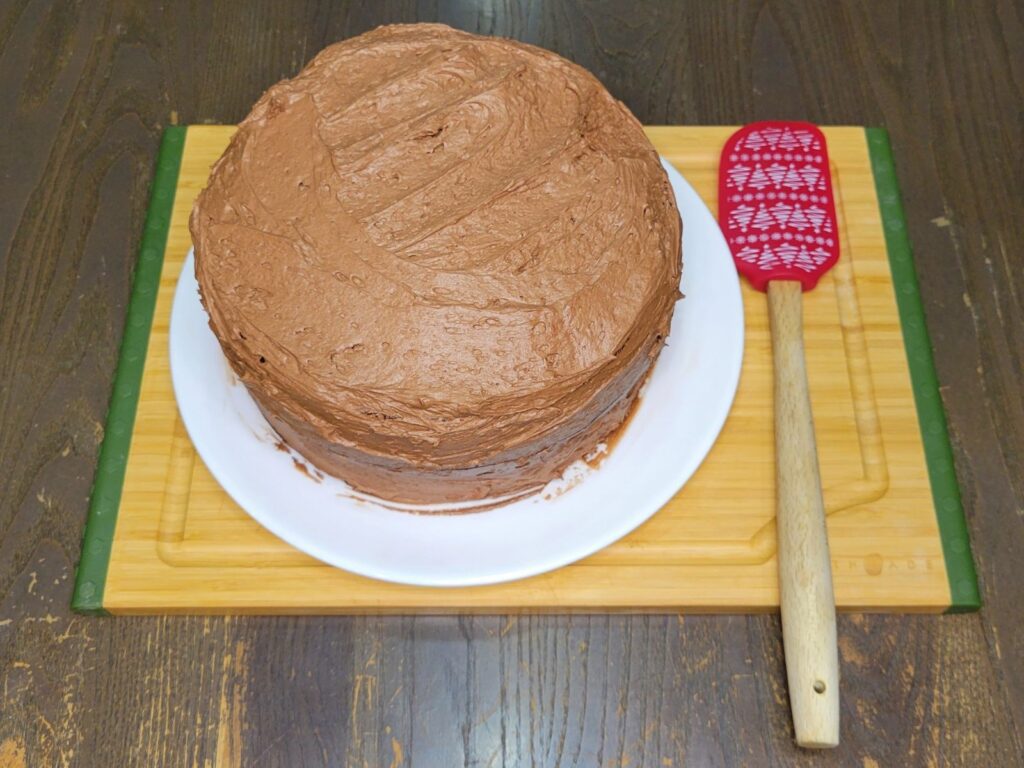 Whole Chocolate cake made from fresh milled wheat. frosted with chocolate buttercream, sitting on a white plate on a bamboo cutting board next to a red spatula