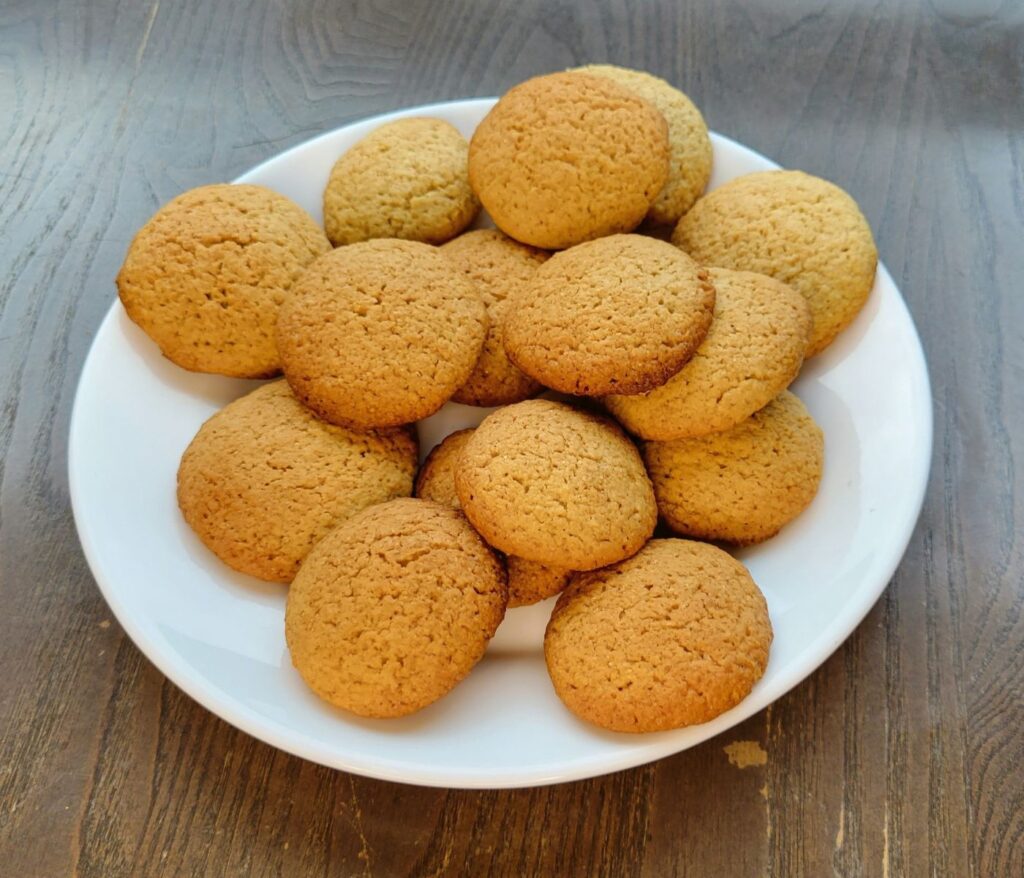 a round white plate on a wooden table that is filled with baked crispy vanilla wafers made with fresh milled flour