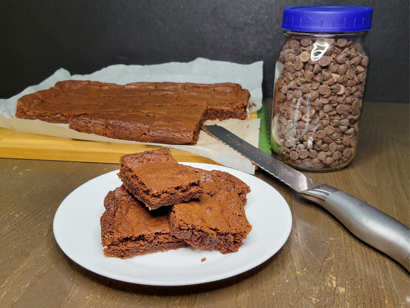 a plate of sliced Brownies made with fresh milled flour in front of a tray of uncut brownies next to a glass mason jar full of chocolate chips.