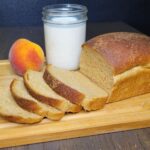 a half sliced loaf of Peaches N' Cream Bread Made With Fresh Milled Flour on a bamboo cutting board next to a glass of milk and a fresh whole peach.