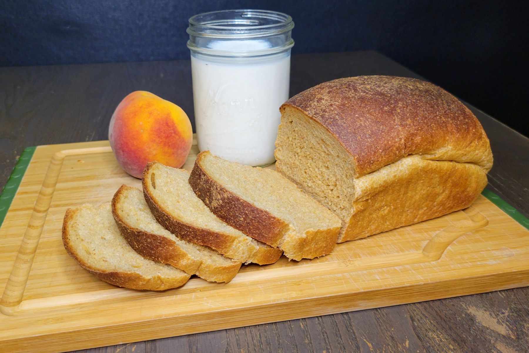a half sliced loaf of Peaches N' Cream Bread Made With Fresh Milled Flour on a bamboo cutting board next to a glass of milk and a fresh whole peach.