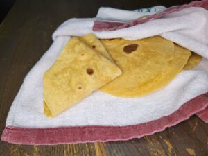 fresh milled flour tortillas wrapped in a kitchen towel, and one is folded many times showing how soft and pliable they are.