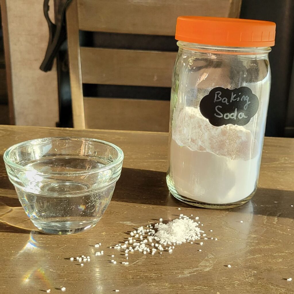 a mason jar of baking soda, glass bowl of water, and a pile of salt on a wooden table.