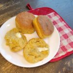 a white plate with 3 fresh milled flour biscuits. one is pulled open showing the inside texture of the biscuit. there is some melted butter on it. and a red and white gingham hot pad underneath.
