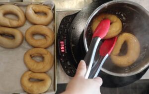 hands using tongs to flip fresh milled flour bagels in the pot of boiling water. boiling the bagels