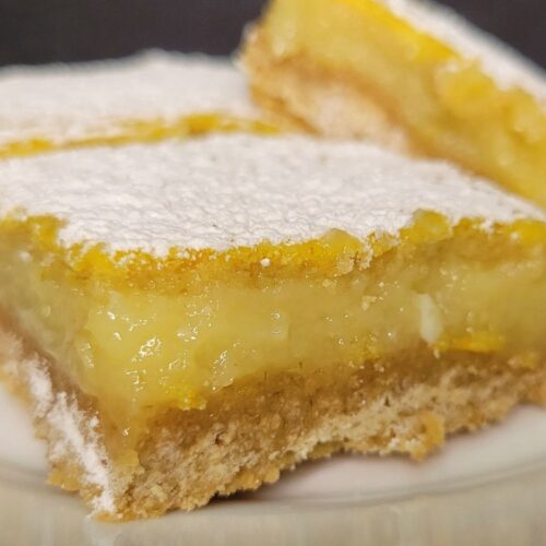a sliced lemon bar made from fresh milled flour on a plate in front of 2 other lemon squares. All dusted with powdered sugar