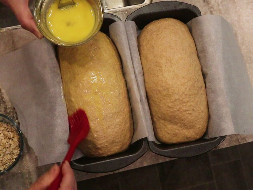 hands using a red pastry brush to brush an egg wash onto the unbaked loafs sitting next to each other getting ready to be baked.