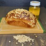 a whole honey oat wheat bread loaf with fresh milled flour sitting on a wooden cutting board. Behind it is a jar of honey and in front a pile of rolled oats.