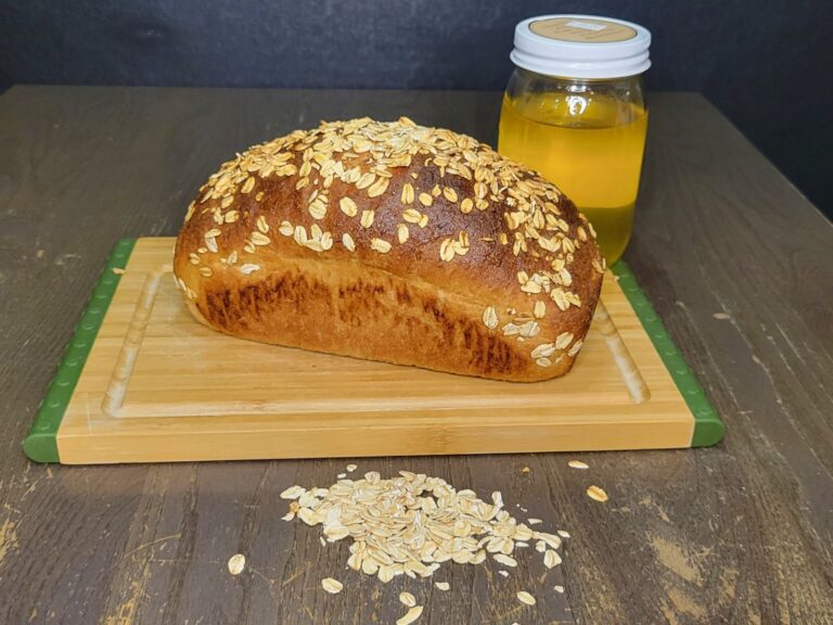 a whole honey oat wheat bread loaf with fresh milled flour sitting on a wooden cutting board. Behind it is a jar of honey and in front a pile of rolled oats.