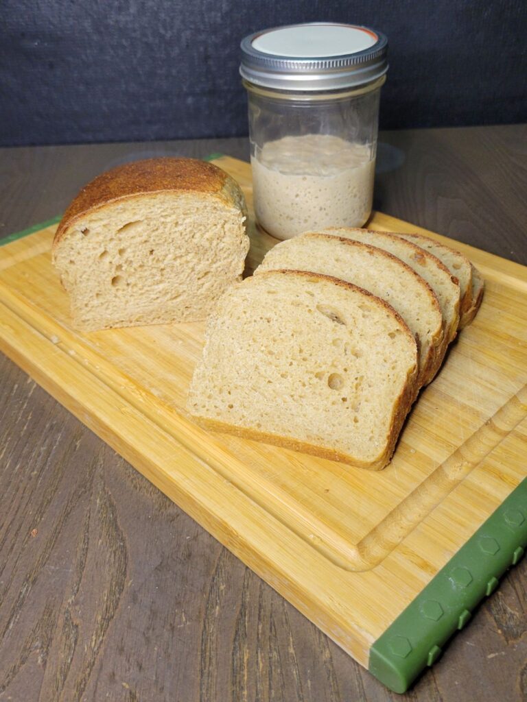 sourdough sandwich bread with fresh milled flour sliced on a cutting board with a jar of sourdough starter behind it