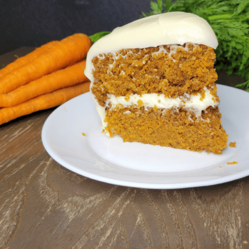 a slice of fresh milled flour carrot cake with cream cheese frosting in front of a bunch of fresh carrots with the green tops still on