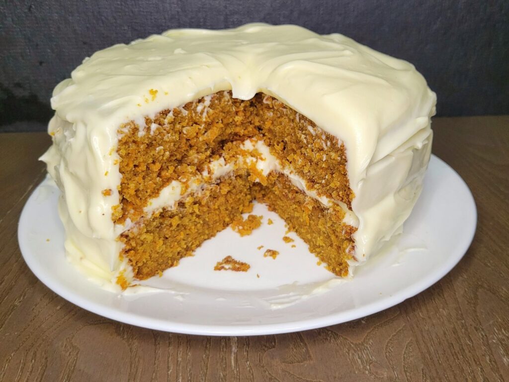 a whole fresh milled flour carrot cake frosted with cream cheese frosting with a slice cut out showing the double layers