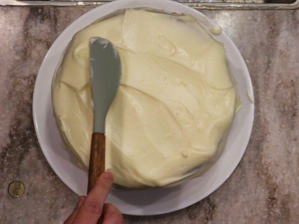 hand icing a carrot cake with cream cheese frosting with a spatula