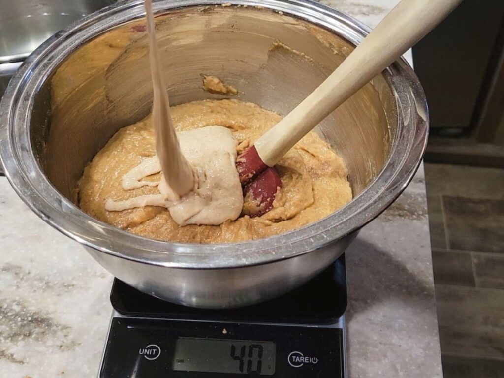 pouring sourdough starter into a bowl of banana bread made with fresh milled flour on a scale.