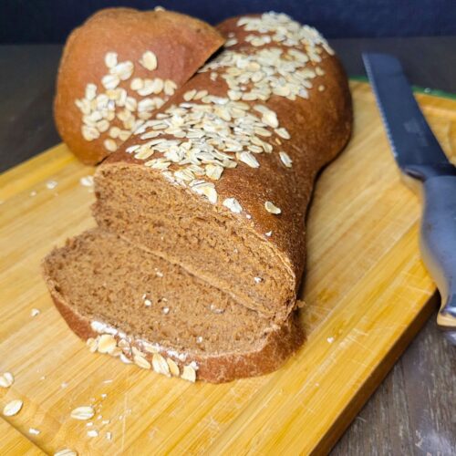 Fresh Milled Flour Brown Bread Loaf topped with rolled oats on a cutting board partially sliced.